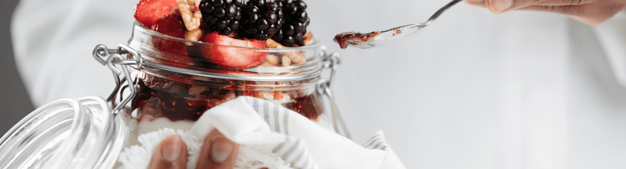 Hands holding open glass jar filled with fresh berries and walnuts on white fabric background