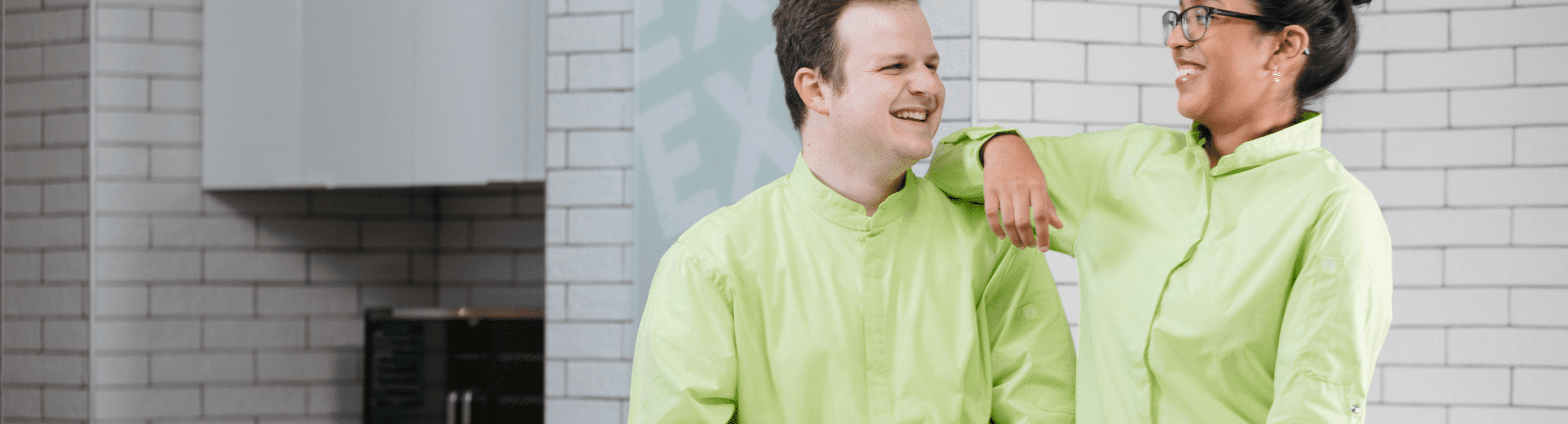Two smiling people wearing green chef coats stand together outside a brick building