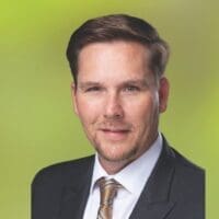 Professional headshot of a smiling businessman in a suit and tie against a green background