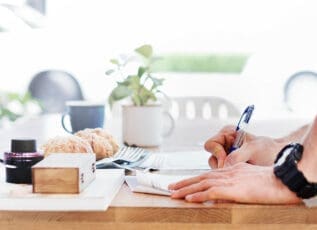 Person writing in notepad at kitchen table with coffee mugs, plant, and pastries