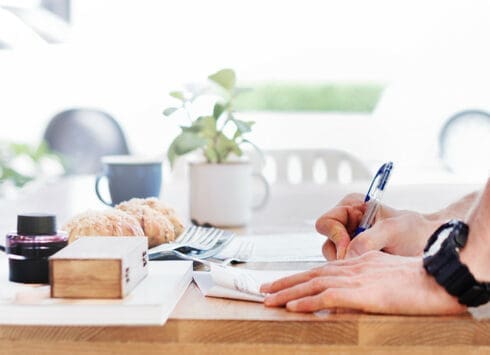 Person writing in notepad at kitchen table with coffee mugs, plant, and pastries