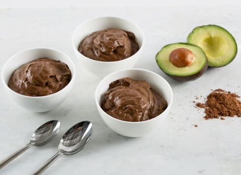 Chocolate avocado mousse in white bowls with avocado halves and cocoa powder on white marble surface