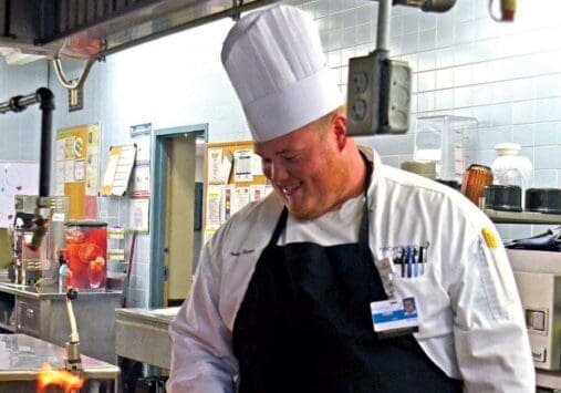 Chef in white uniform and hat smiling while cooking in industrial kitchen