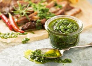 Chimichurri sauce in glass jar with fresh herbs and red chili peppers on rustic wooden board