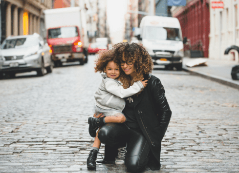 Smiling woman hugging young girl on city street surrounded by cars and buildings