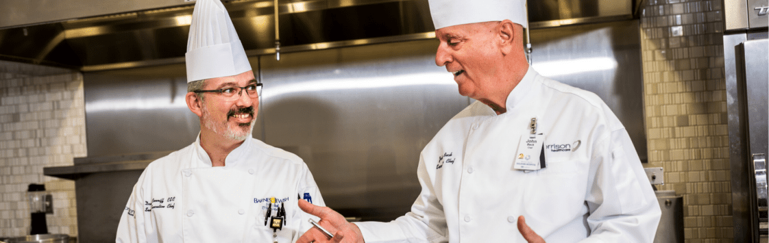 Two chefs in white uniforms and hats smiling and talking in a commercial kitchen