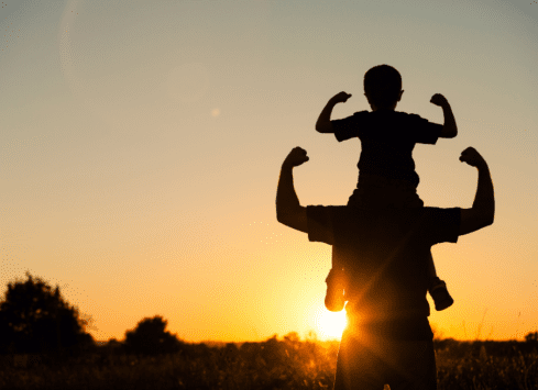 Silhouette of child flexing muscles on father's shoulders at sunset in field