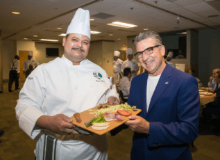 Morrison Healthcare chefs presenting healthy meal options at an event, smiling and holding a cutting board with vegetables.