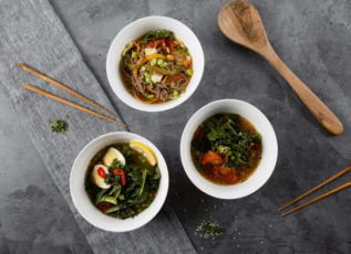 Vegan noodle bowls with vegetables and greens, served with wooden utensils on a gray stone surface