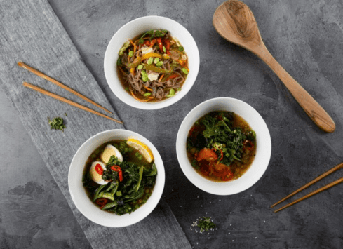 Vegan noodle bowls with vegetables and greens, served with wooden utensils on a gray stone surface