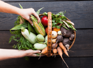 Basket of fresh vegetables including corn, tomato, carrots, herbs held by hands on wooden table