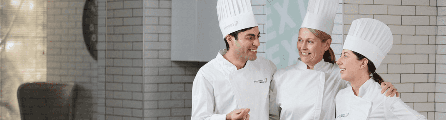 Three smiling chefs in white uniform standing together in a commercial kitchen