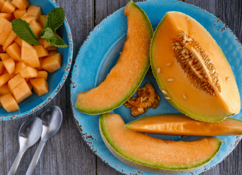 Cantaloupe melon slices on blue plate with diced cantaloupe in bowl and spoons on rustic wood table
