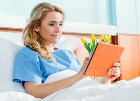 Woman in hospital bed reading book next to vase of yellow tulips