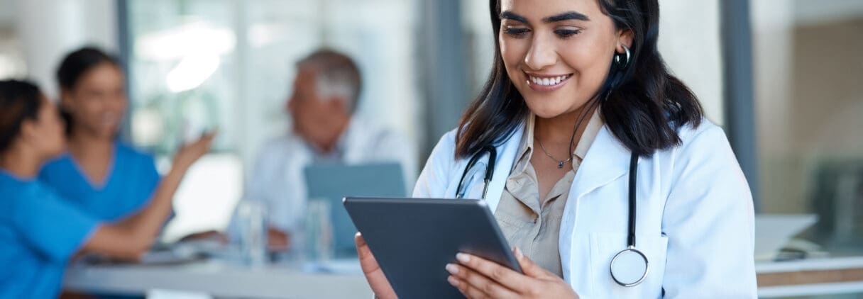 Smiling female healthcare professional holding tablet in medical office with colleagues in background