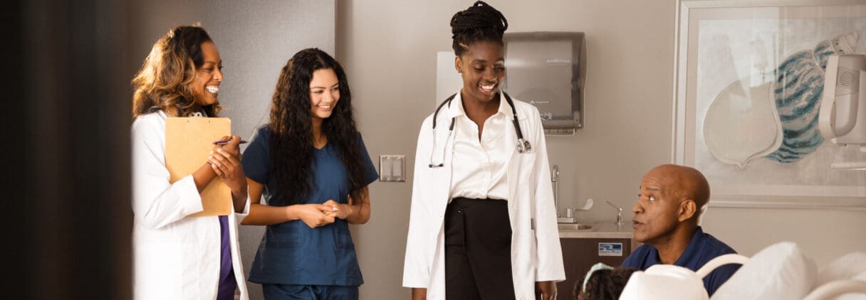 Three friendly healthcare professionals talk with a smiling patient in a hospital exam room
