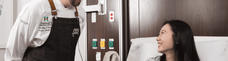 Morrison Healthcare employee in chef uniform talking with patient in hospital room
