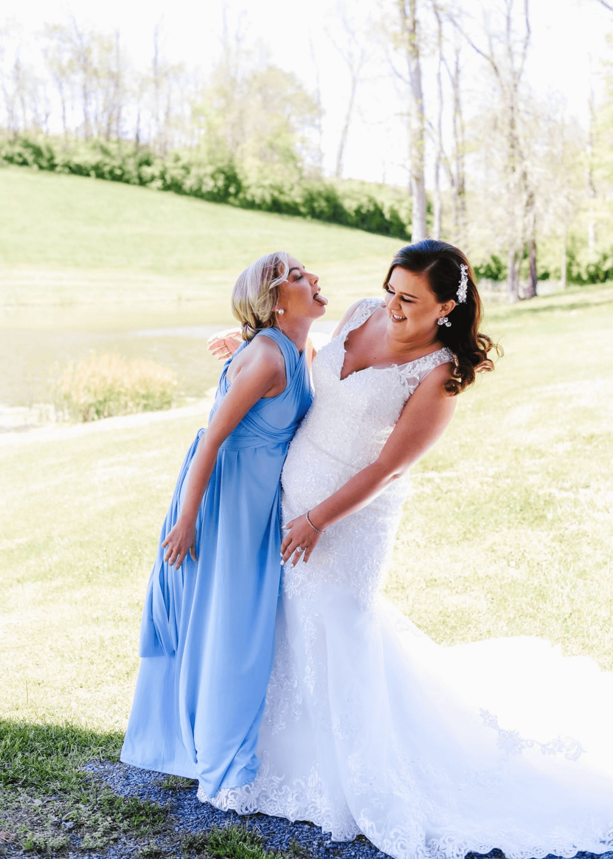 Bride in blue dress and bridesmaid laughing together on grass path surrounded by trees