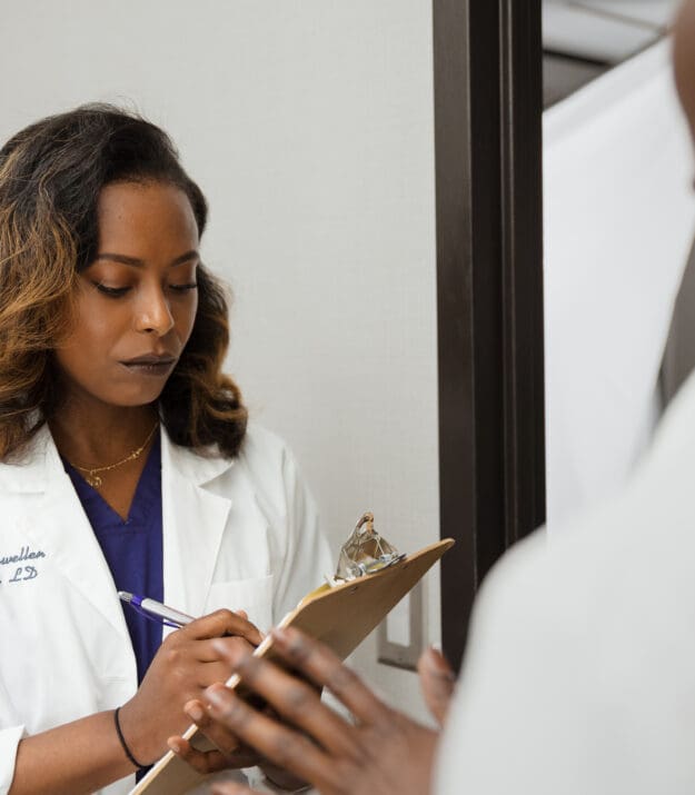 Morrison Healthcare clinician in a white coat carefully writing on a clipboard in a hospital hallway while conferring with a colleague, illustrating focused professional competency.