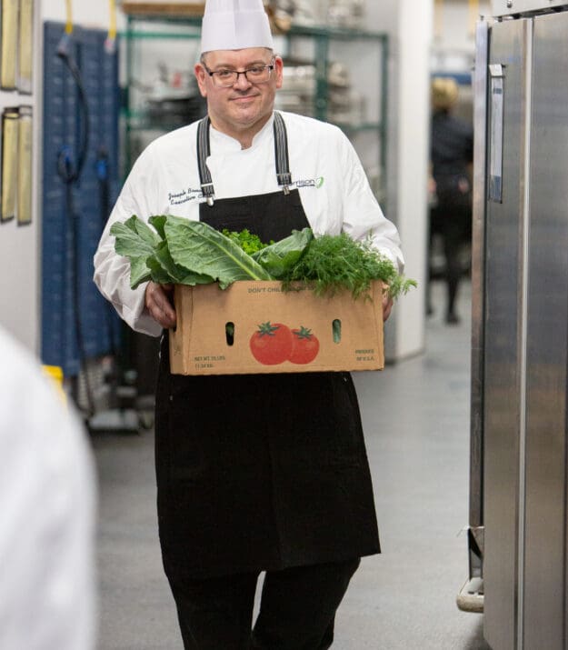 Chef in a Morrison Healthcare kitchen holding a cardboard box filled with fresh leafy greens and herbs.